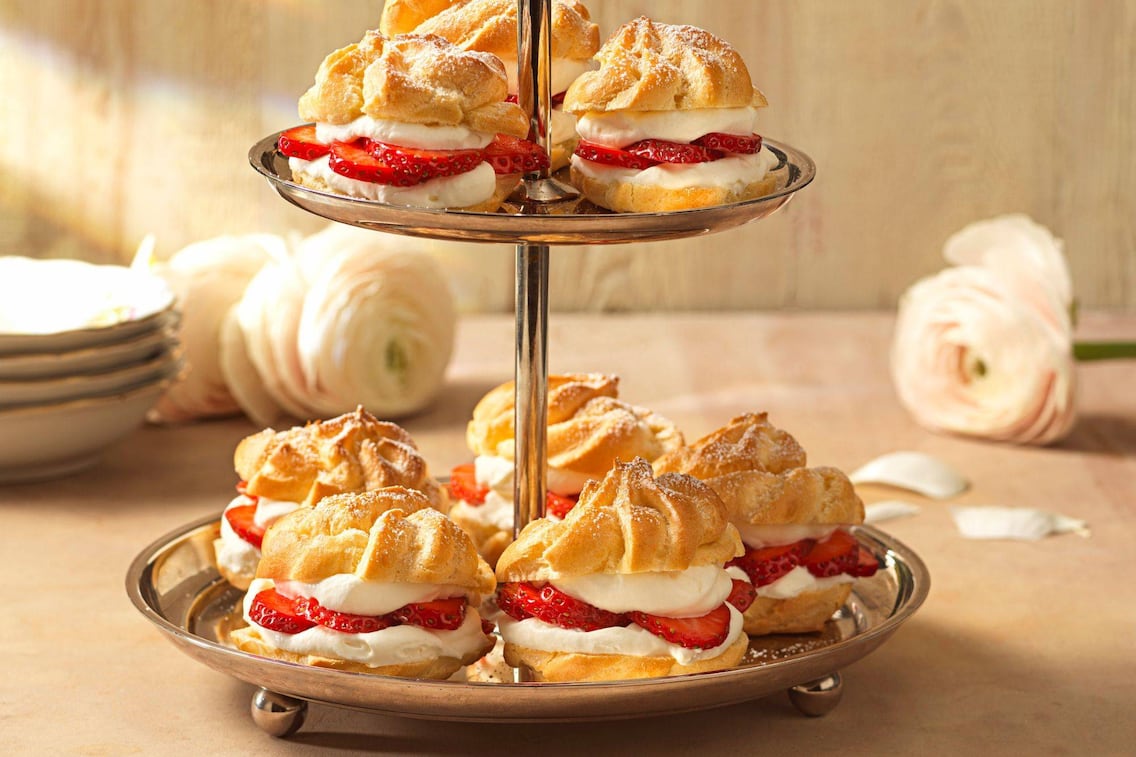Brandteigkrapferl mit Schlagobers und Erdbeerscheiben gefüllt und auf einer silbernen Etagere fotografiert.