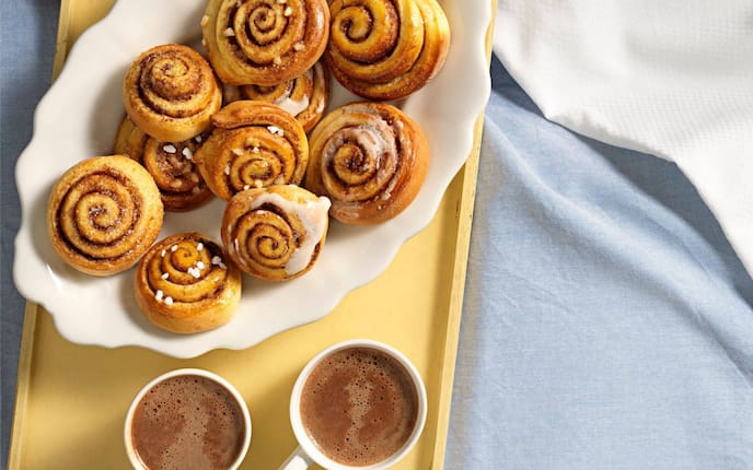 Kleine Zimtstnecken mit Hagelzucker oder Zuckerguss auf einer weißen Keramikservierplatte angerichtet. Die Platte steht auf einem alten gelben Holztablett mit zwei Häferl Kakao und einem hellblauen Tischtuch