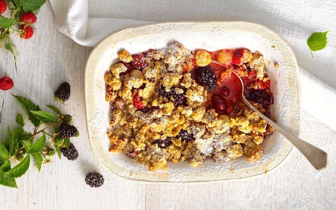Frische Brombeeren, Erdbeeren, Ribisel in einem tiefen Keramikschüsserl mit Streusel überbacken und Staubzucker bestreut. Alles steht auf einem weißen Tisch, von oben fotografiert, daneben liegt ein Erdbeerzweig und ein Brombeerzweig.