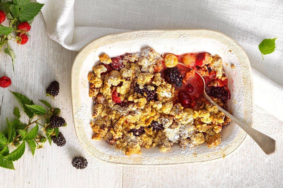 Frische Brombeeren, Erdbeeren, Ribisel in einem tiefen Keramikschüsserl mit Streusel überbacken und Staubzucker bestreut. Alles steht auf einem weißen Tisch, von oben fotografiert, daneben liegt ein Erdbeerzweig und ein Brombeerzweig.