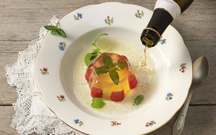 Ein Wackelpudding mit ganzen Himbeeren und Minze in einem tiefen Suppenteller mit Blumenmuster angerichtet, ein Schuss Sekt wird gerade dazugeleert.