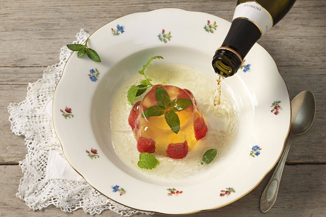 Ein Wackelpudding mit ganzen Himbeeren und Minze in einem tiefen Suppenteller mit Blumenmuster angerichtet, ein Schuss Sekt wird gerade dazugeleert.