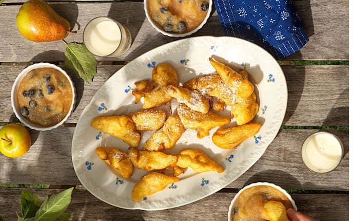 Holztisch von oben fotografiert, darauf steht ein Serviertablett aus Keramik in weiß mit kleinen blauen Blümchen (Vergissmeinnicht) darauf liegen gebackene Apfel- und Birnenspalten mit Staubzucker bestreut. Daneben stehen drei Schälchen Mus mit Schwarzbeeren. und zwei Gläser Milch.