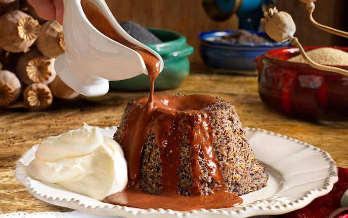 Mohnküchlein, wird aus einer kleinen Keramiksauciere mit Schokoladensauce übergossen. Das Küchlein steht au feinem Keramikteller, daneben liegt noch ein Tupfer Schlagobers.