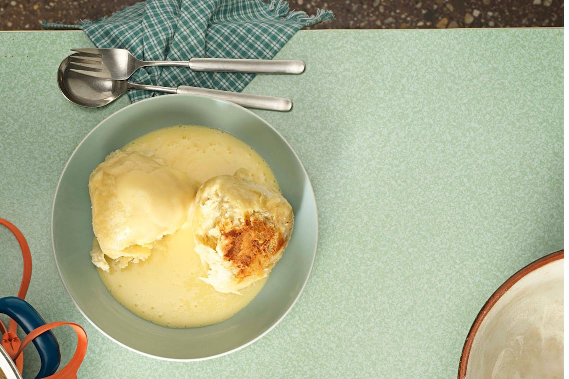 Dampfnudeln in Vanillesauce in einem tiefen Teller angerichtet auf einem alten Omatisch mit Besteck und karierter Serviette
