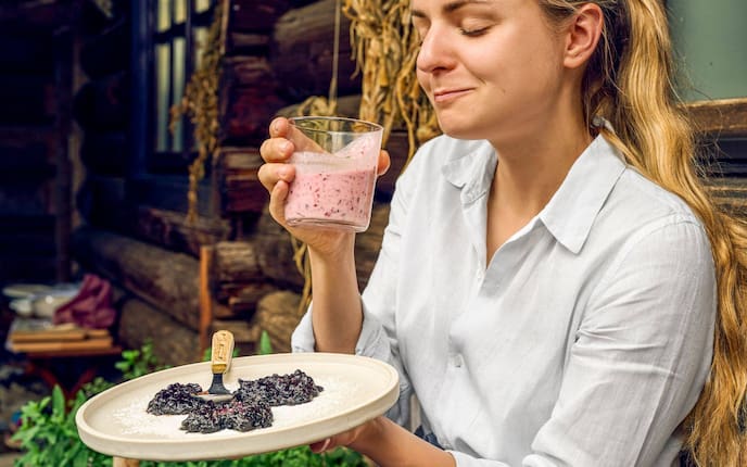 Paula Bründl genießt vor einer Holzbauernhütte auf einer Bank einen Schluck Buttermilch aus einem Glas und hat dabei genussvoll die Augen geschlossen. In der Hand hält sie ienen Teller mit drei Stück Moosbeernocken.
