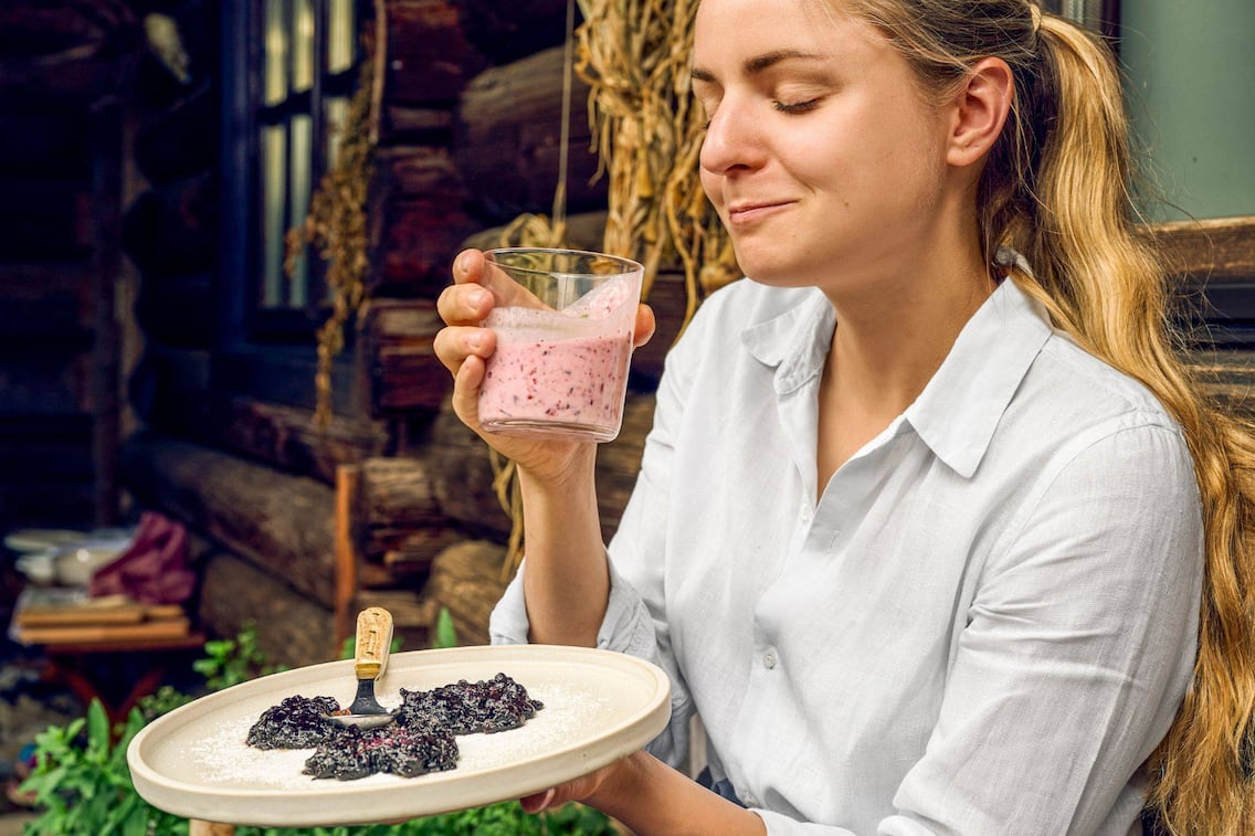 Paula Bründl genießt vor einer Holzbauernhütte auf einer Bank einen Schluck Buttermilch aus einem Glas und hat dabei genussvoll die Augen geschlossen. In der Hand hält sie ienen Teller mit drei Stück Moosbeernocken.