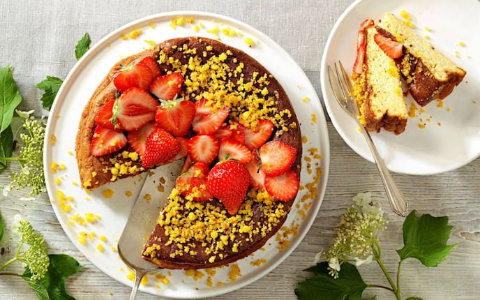 Ein ganzer Kuchen auf einem weißen Teller mit Riebelstreuseln und halbierten frischen Erdbeeren. Daneben steht ein Dessertteller mit zwei Stücke vom Kuchen und einer Dessertgabel.