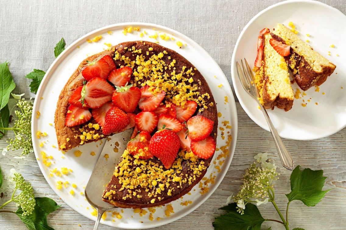 Ein ganzer Kuchen auf einem weißen Teller mit Riebelstreuseln und halbierten frischen Erdbeeren. Daneben steht ein Dessertteller mit zwei Stücke vom Kuchen und einer Dessertgabel.