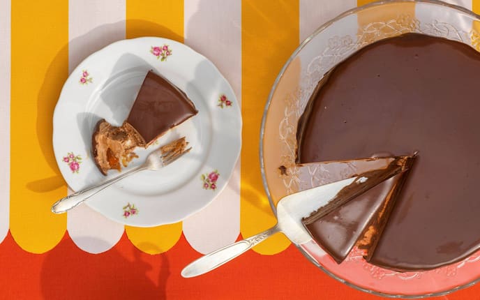 Eine Eistorte, die wie eine Sachertorte aussieht, steht auf einem Tortenservierteller aus Glas, ein Stück ist runtergeschnitten und liegt daneben auf einem Dessertteller mit vintage Blumenmuster. Ein weiteres Stück ist heruntergeschnitten und liegt auf dem Tortenteller auf dem Tortenheber.