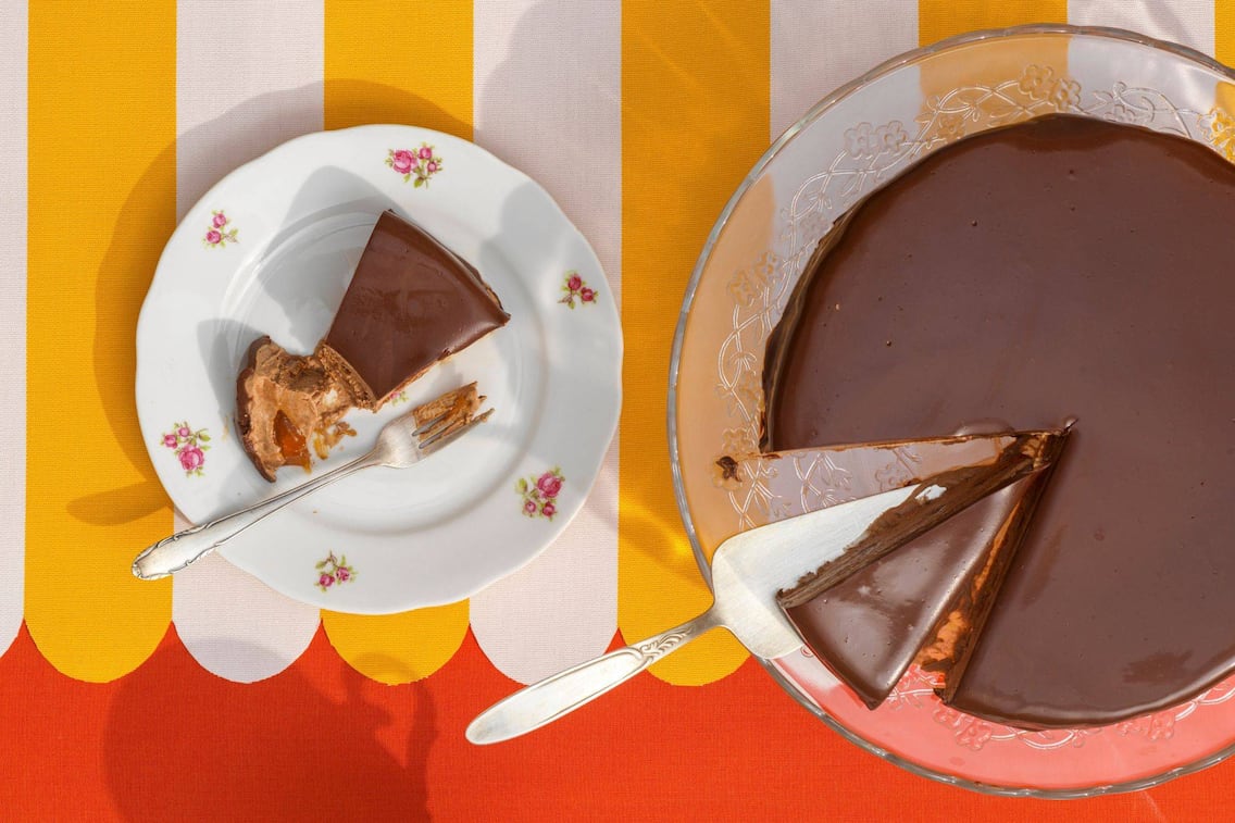 Eine Eistorte, die wie eine Sachertorte aussieht, steht auf einem Tortenservierteller aus Glas, ein Stück ist runtergeschnitten und liegt daneben auf einem Dessertteller mit vintage Blumenmuster. Ein weiteres Stück ist heruntergeschnitten und liegt auf dem Tortenteller auf dem Tortenheber.