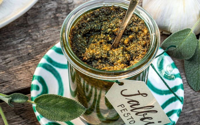 Salbeipesto in einem Rexglas. Darin steckt ein Löffel. Das Glas steht auf einem Gmundner Keramikteller, grün geflammt. Daneben lieben ein paar Salbeiblätter. Um das Glas ist ein Papieretikett mit grüner Schnur befestigt. Darauf steht: Salbei Pesto. Alles steht auf einem alten Holztisch.