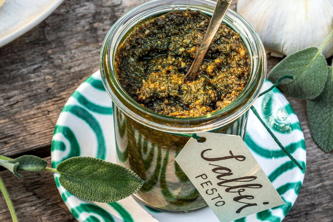 Salbeipesto in einem Rexglas. Darin steckt ein Löffel. Das Glas steht auf einem Gmundner Keramikteller, grün geflammt. Daneben lieben ein paar Salbeiblätter. Um das Glas ist ein Papieretikett mit grüner Schnur befestigt. Darauf steht: Salbei Pesto. Alles steht auf einem alten Holztisch.