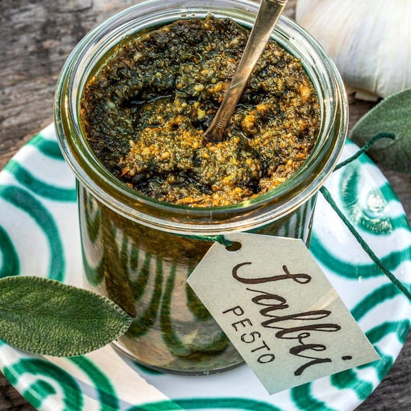 Salbeipesto in einem Rexglas. Darin steckt ein Löffel. Das Glas steht auf einem Gmundner Keramikteller, grün geflammt. Daneben lieben ein paar Salbeiblätter. Um das Glas ist ein Papieretikett mit grüner Schnur befestigt. Darauf steht: Salbei Pesto. Alles steht auf einem alten Holztisch.