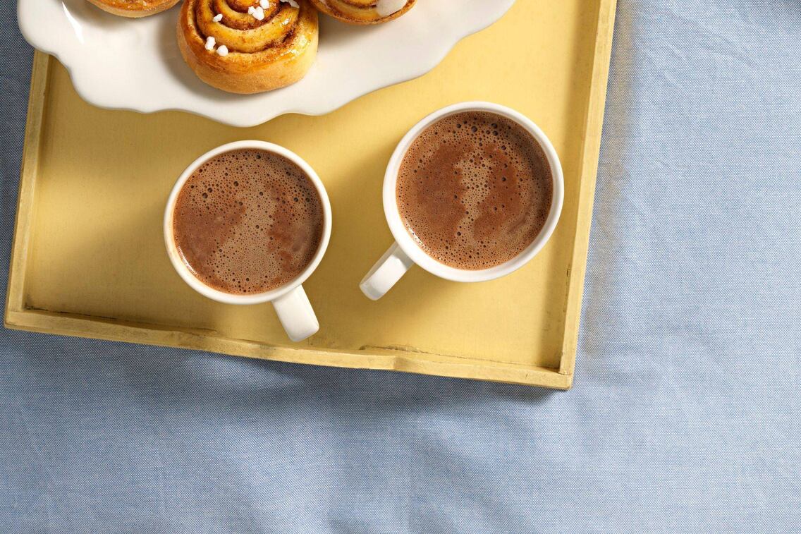 Zwei weiße Häferl mit Kakao, stehen auf einem alten gelben Holztablett, daneben kann man angeschnitte kleine Zimtschnecken sehen. Das Tablett steht auf einem hellblauen Tischtuch