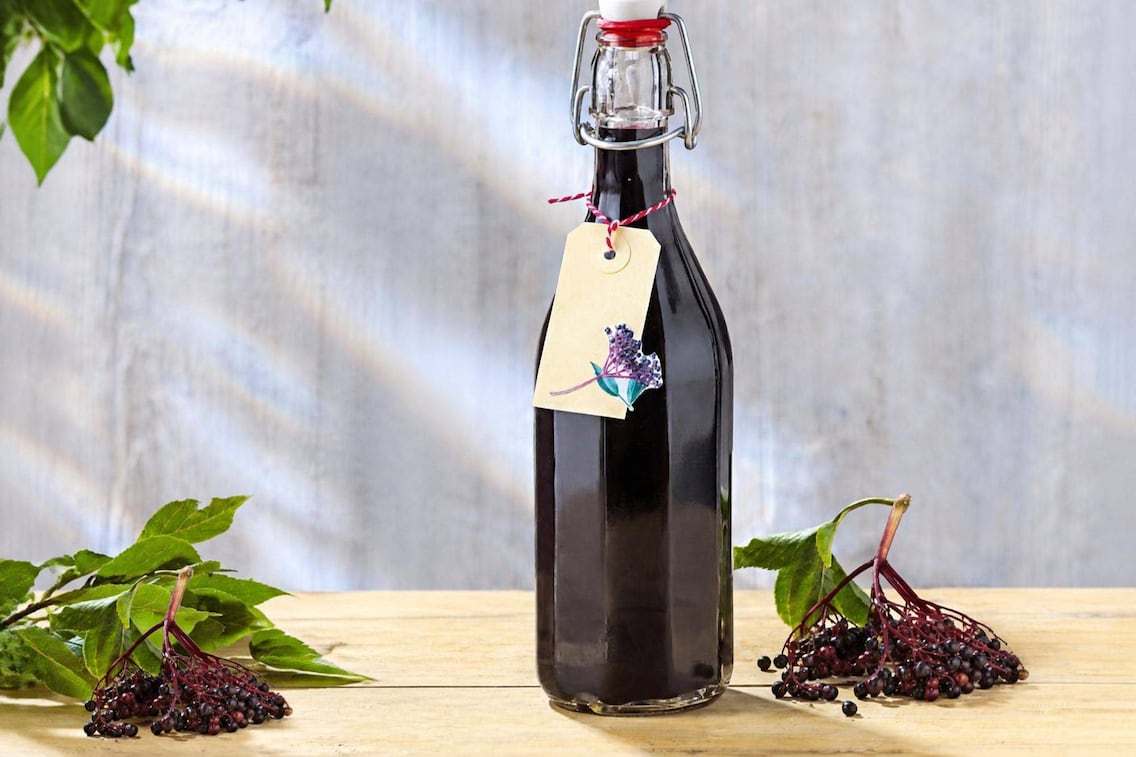 Selbstgemachter Hollerbeerensirup in einer Bügelverschlussflasche mit weißem Keramikverschluss und rotem Garn. Ein Kraftpapier-Anhänger mit Hollerbeeren-Illustration hängt am Flaschenhals. Links und rechts liegen frische schwarze Hollerbeeren-Rispen mit Blättern auf einem hellen Holztisch.