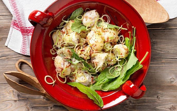 Kleine gekochte Erdäpfel mit Lauchringen und Sauerampfer, Marinade und Speck in einer roten Keramikschüssel mit Henkel. Daneben liegt ein alter Mehllöffel und ein Salatbesteck aus Holz. Alles steht auf einem Holztisch und ein Tischtuch in natur mit roter Bordüre liegt teilweise unter der roten Salatschüssel.