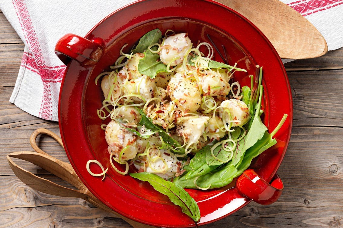 Kleine gekochte Erdäpfel mit Lauchringen und Sauerampfer, Marinade und Speck in einer roten Keramikschüssel mit Henkel. Daneben liegt ein alter Mehllöffel und ein Salatbesteck aus Holz. Alles steht auf einem Holztisch und ein Tischtuch in natur mit roter Bordüre liegt teilweise unter der roten Salatschüssel.