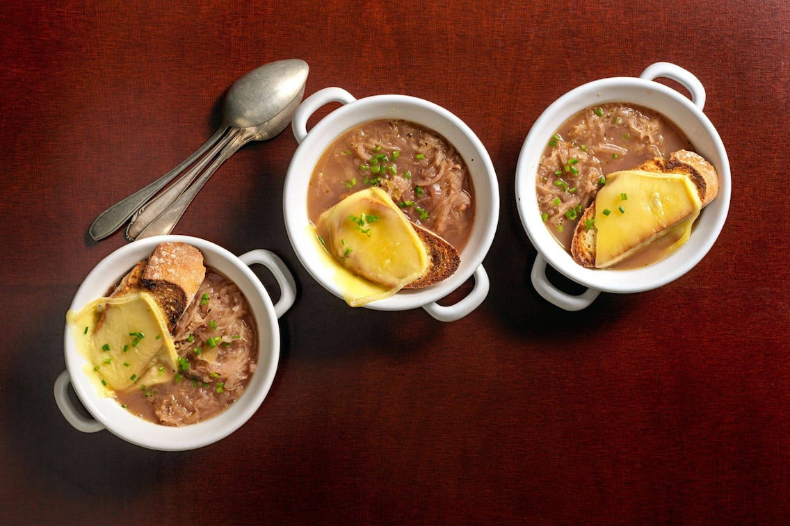 Sauerkrautsuppe in drei weißen Keramiksuppenschüsseln angerichtet, darauf liegt eine Scheibe knuspriges Weißbrot mit überbackenem Käse. Daneben liegen drei alte Löffel übereinandergelegt auf einem roten Untergrund.