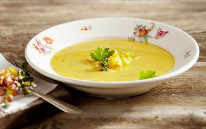 ein blumiger Keramikteller mit einer Erdäpfelsuppe, daneben liegt auf einem Holzbrett angeschnitten gewürfelter Speck