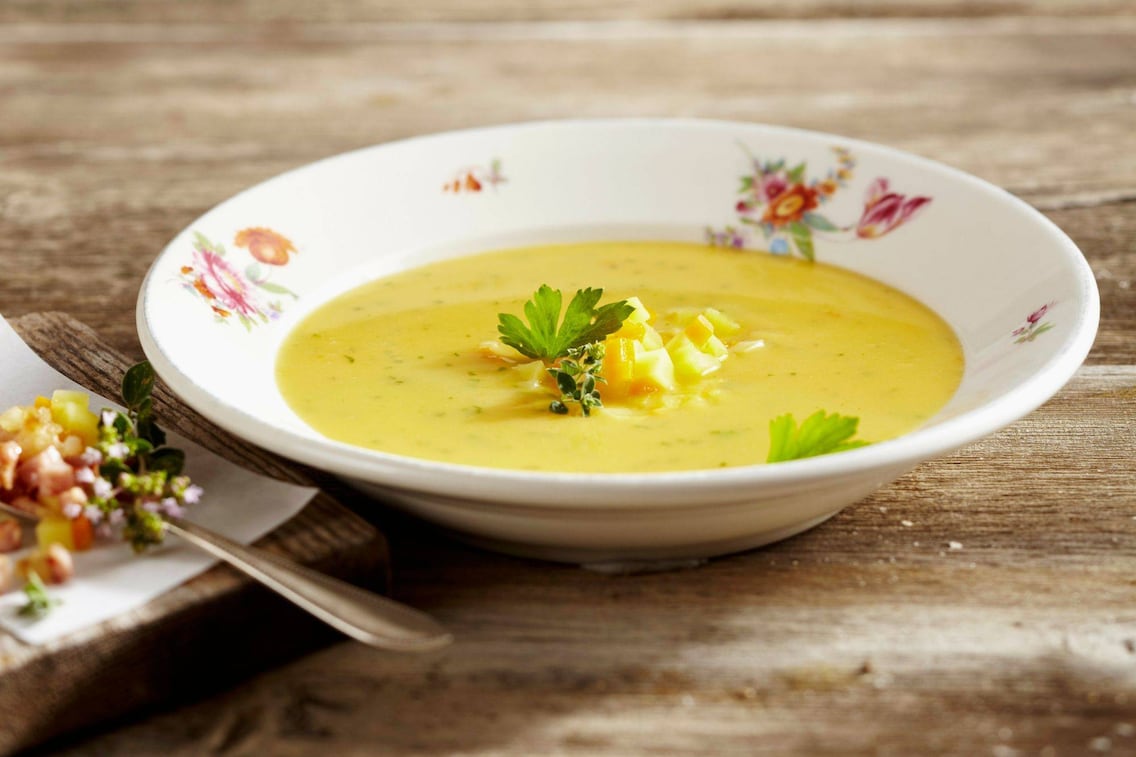 ein blumiger Keramikteller mit einer Erdäpfelsuppe, daneben liegt auf einem Holzbrett angeschnitten gewürfelter Speck