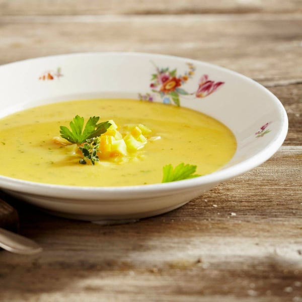 ein blumiger Keramikteller mit einer Erdäpfelsuppe, daneben liegt auf einem Holzbrett angeschnitten gewürfelter Speck