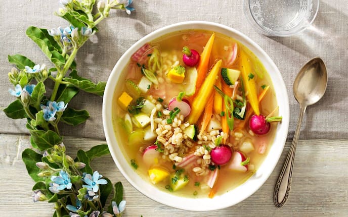 Karotten, Radischen, Zuchhini, Rollgerste in einer Gemüsesuppe. Daneben liegt ein Suppenlöffel und ein Gals perlendes Mineralwasser. Alles steht auf einer Stofftischdecke in natur.