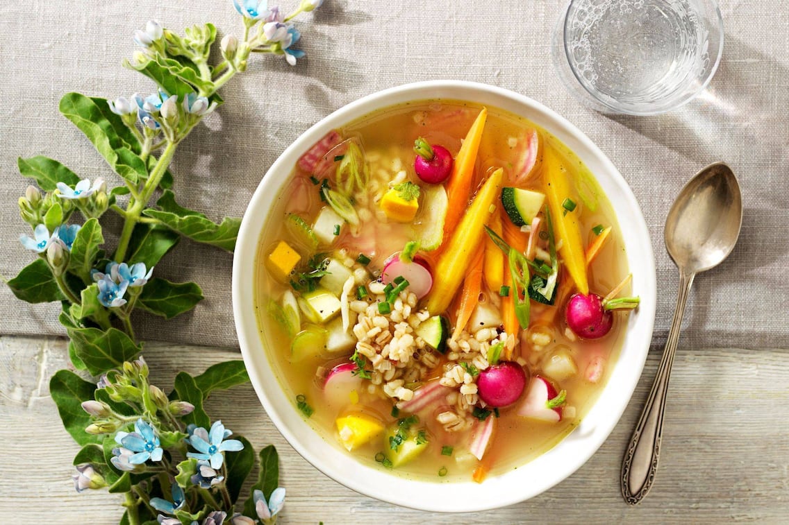 Karotten, Radischen, Zuchhini, Rollgerste in einer Gemüsesuppe. Daneben liegt ein Suppenlöffel und ein Gals perlendes Mineralwasser. Alles steht auf einer Stofftischdecke in natur.