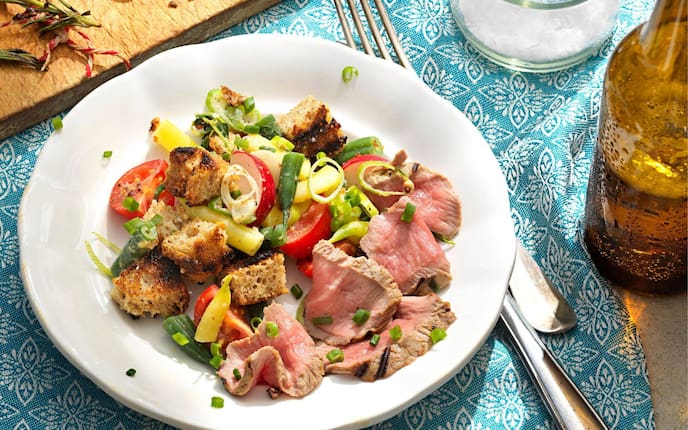 Weißer, geschwungener Teller mit gegrillten Brotwürfeln, Tomaten, Radieschen, Fisolen und aufgeschnittenem Lammfleisch. Daneben steht eine Flasceh Bier, und Besteck liegt neben dem Teller. Alles steht auf einer türkisen Stischdecke mit weißem Stickmuster.