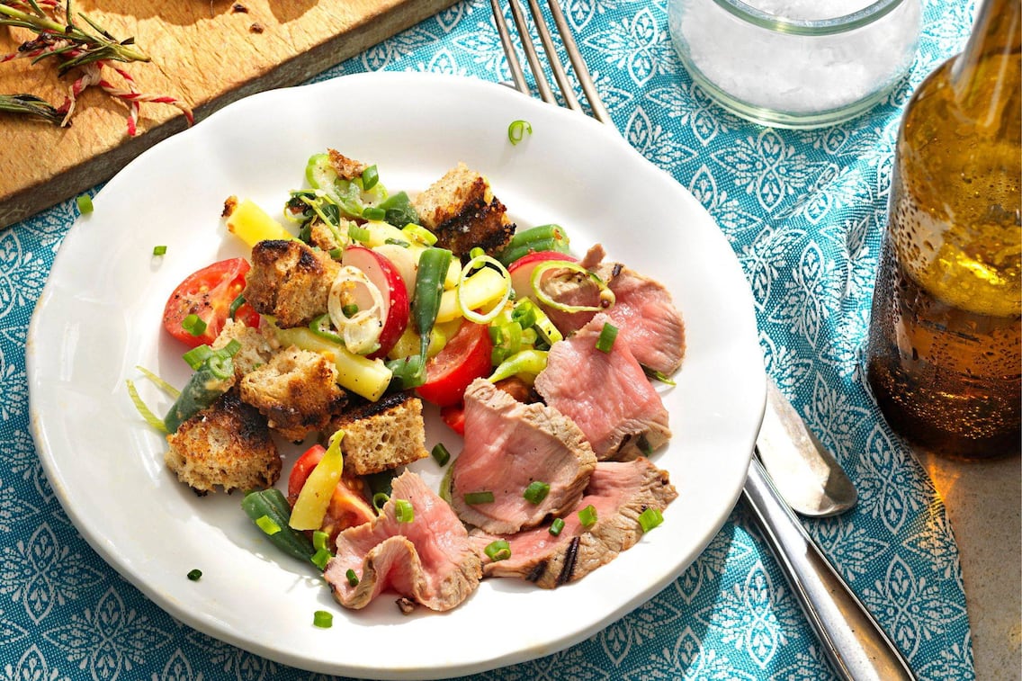 Weißer, geschwungener Teller mit gegrillten Brotwürfeln, Tomaten, Radieschen, Fisolen und aufgeschnittenem Lammfleisch. Daneben steht eine Flasceh Bier, und Besteck liegt neben dem Teller. Alles steht auf einer türkisen Stischdecke mit weißem Stickmuster.