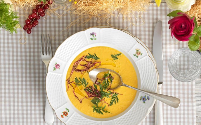 Karottensuppe in einem Suppenteller mit Blumenmuster und Goldrand, in der Suppe schwiemmen Kräuter.