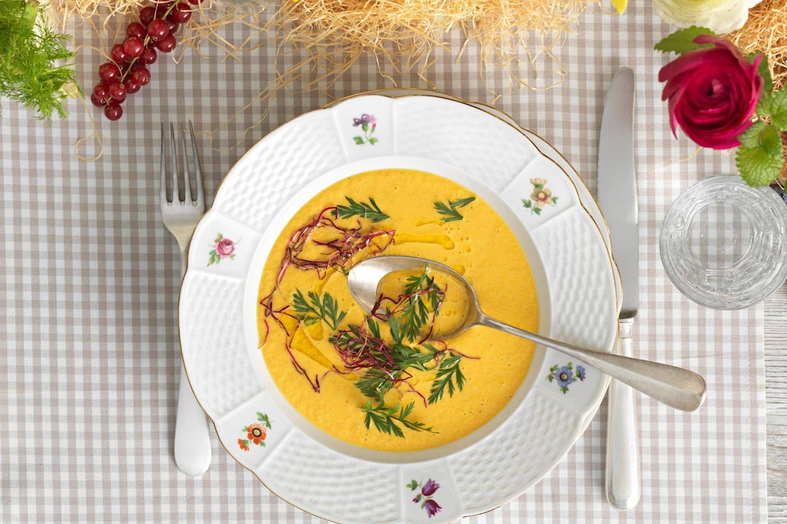 Karottensuppe in einem Suppenteller mit Blumenmuster und Goldrand, in der Suppe schwiemmen Kräuter.