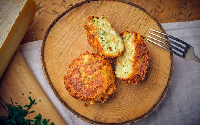 Ein Kaspressknödel und ein zweiter aufgeschnittener auf einer Holzscheibe angerichtet. Daneben liegt ein Käselaib.