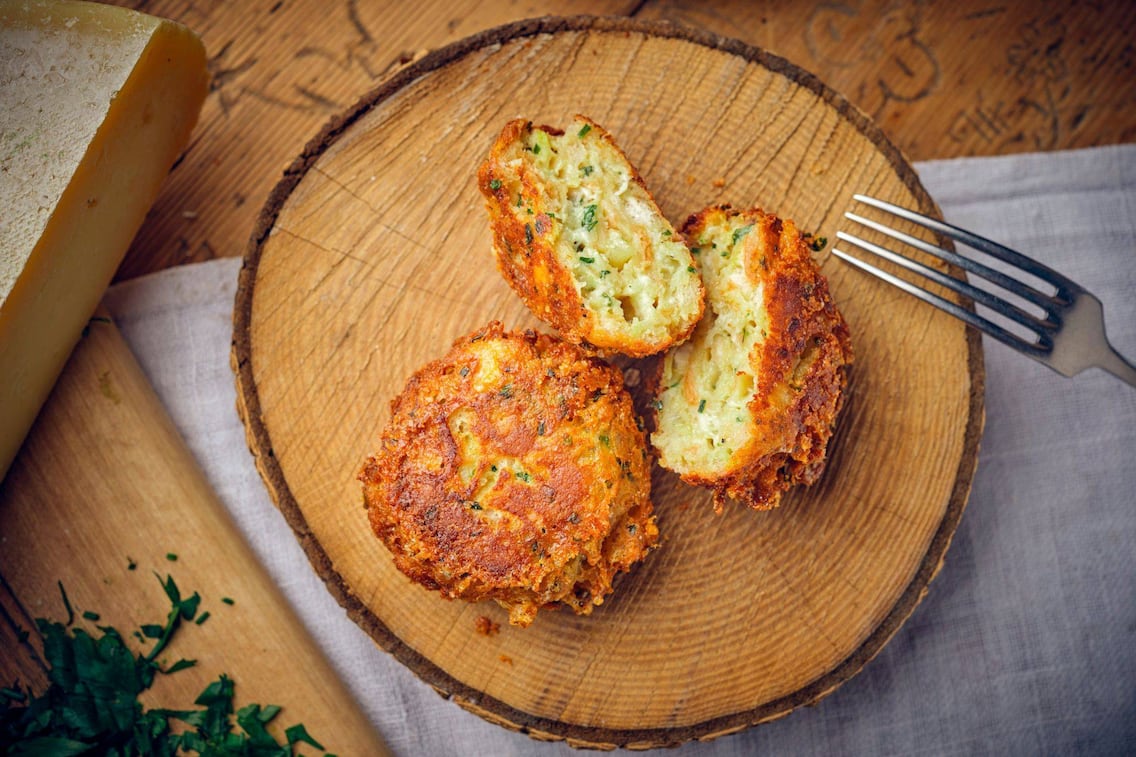 Ein Kaspressknödel und ein zweiter aufgeschnittener auf einer Holzscheibe angerichtet. Daneben liegt ein Käselaib.