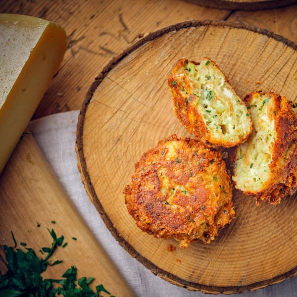 Ein Kaspressknödel und ein zweiter aufgeschnittener auf einer Holzscheibe angerichtet. Daneben liegt ein Käselaib.