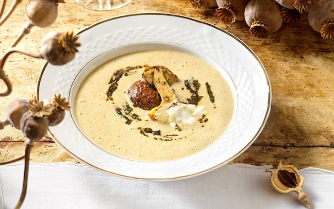 Steinpilzcremesuppe mit zwei angebratenen Steinpilzhälften und Mohn und Schlagobers in einem tiefen weißen Teller mit Goldrand serviert. Der Teller steht auf einem alten Holzuntergrund und getrockneter Mohn liegt als Deko daneben.