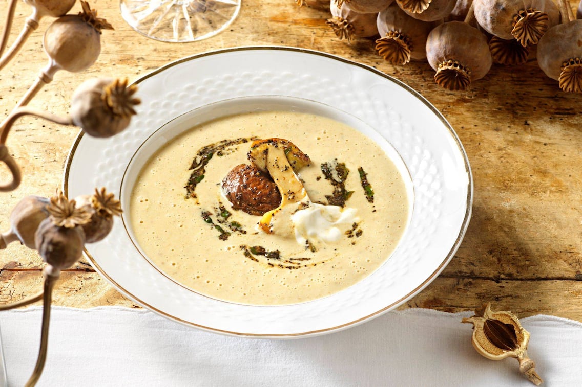 Steinpilzcremesuppe mit zwei angebratenen Steinpilzhälften und Mohn und Schlagobers in einem tiefen weißen Teller mit Goldrand serviert. Der Teller steht auf einem alten Holzuntergrund und getrockneter Mohn liegt als Deko daneben.