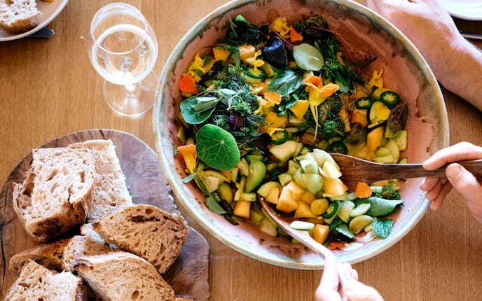 Gurken-Melonen-Salat frisch angerichtet in einer großen Keramikschüssel, in der gerade jemand mit Salatbesteck umrührt. Daneben steht ein Teller mit frisch geschnittenem Brot.