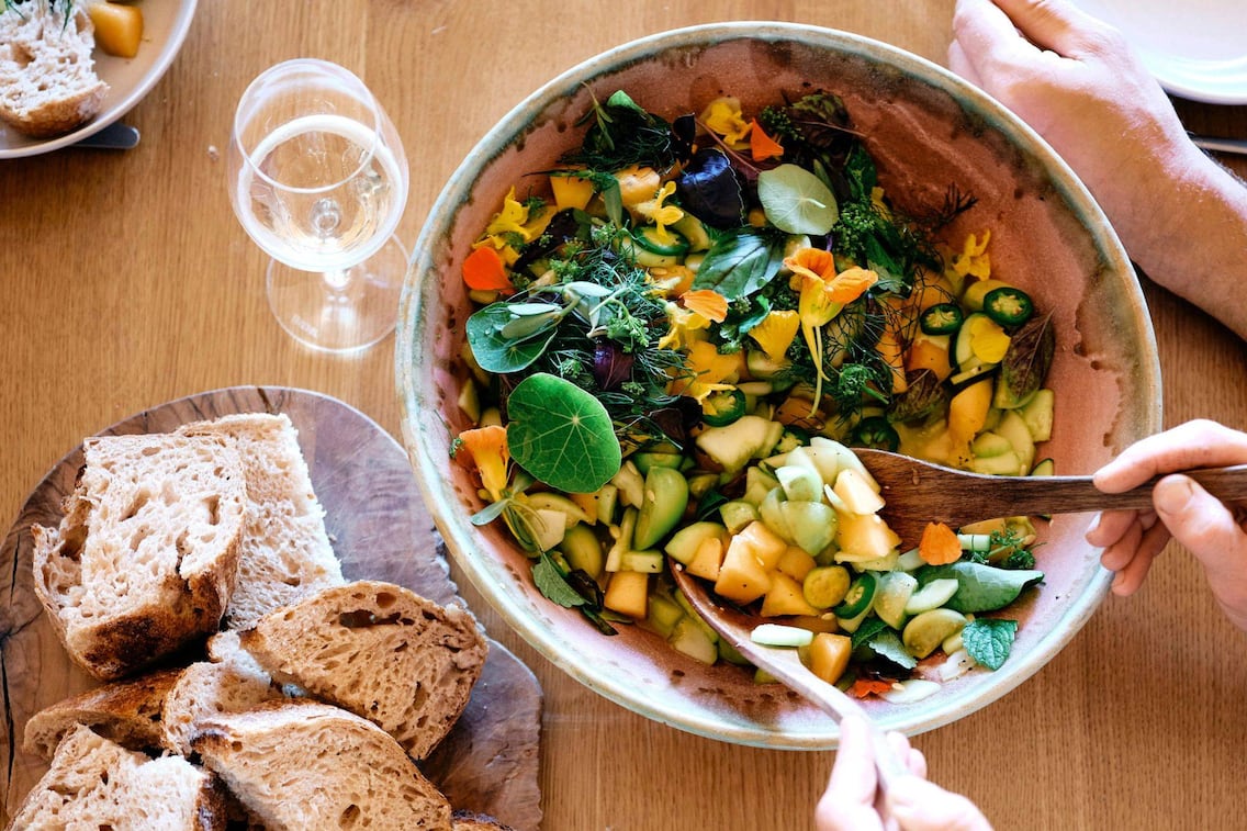 Gurken-Melonen-Salat frisch angerichtet in einer großen Keramikschüssel, in der gerade jemand mit Salatbesteck umrührt. Daneben steht ein Teller mit frisch geschnittenem Brot.