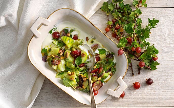 Stachelbeeren, Gurken, Weintrauben und Kräuter in einem Salat mit Dressing vermischt. Der Salat ist in einer tiefen Servierschüssel mit Goldrand angerichtet. Es liegt ein Esslöffel auf dem Salat. Daneben liegt ein Zweig mit Stachelbeeren. Alles zusammen steht auf einem weißen Holztisch.