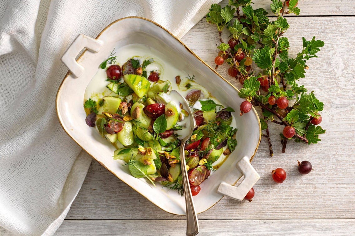 Stachelbeeren, Gurken, Weintrauben und Kräuter in einem Salat mit Dressing vermischt. Der Salat ist in einer tiefen Servierschüssel mit Goldrand angerichtet. Es liegt ein Esslöffel auf dem Salat. Daneben liegt ein Zweig mit Stachelbeeren. Alles zusammen steht auf einem weißen Holztisch.