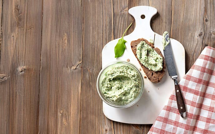 Aufstrich mit Sauerampfer in einer kleinen Glaschüssel auf einem weißen Brett. Daneben liegt eine Scheibe Brot mit Aufstrich und ein Buttermesser. Daneben liegt ein rot-weiß kariertes Geschirrtuch. Alles steht auf einem Holztisch.