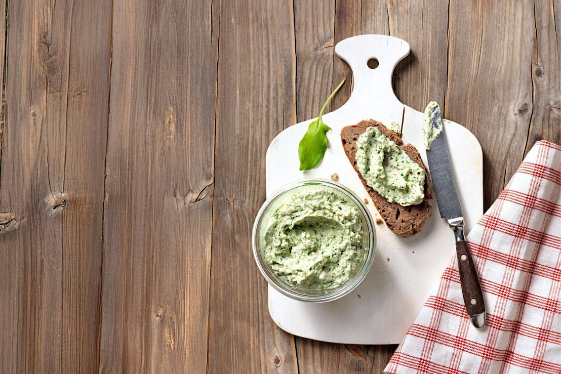 Aufstrich mit Sauerampfer in einer kleinen Glaschüssel auf einem weißen Brett. Daneben liegt eine Scheibe Brot mit Aufstrich und ein Buttermesser. Daneben liegt ein rot-weiß kariertes Geschirrtuch. Alles steht auf einem Holztisch.