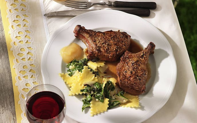 Wildschweinkoteletts mit Mangoldfleckerln auf einem weißen Teller angerichtet. Er steht auf einer weißen Serviette, darunter liegt noch eine gelbe, die hervorblitzt. Alles steht auf einem alten Holztisch von oben fotografiert, mit einem Glas Rotwein und einem Glasschüsserl mit Apfelmus.