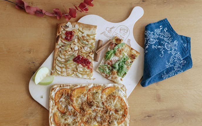 Drei Flammkuchen auf einem Holzbrett der Marke Tante Fanny: mit Äpfeln und Mandelblättchen, mit Rucola und Speck sowie mit gelb-orangefarbenen Tomaten und Käse.