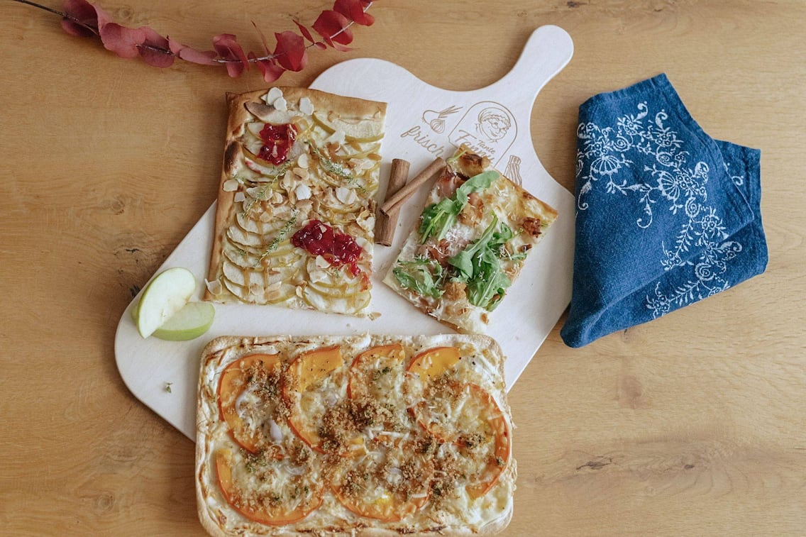 Drei Flammkuchen auf einem Holzbrett der Marke Tante Fanny: mit Äpfeln und Mandelblättchen, mit Rucola und Speck sowie mit gelb-orangefarbenen Tomaten und Käse.